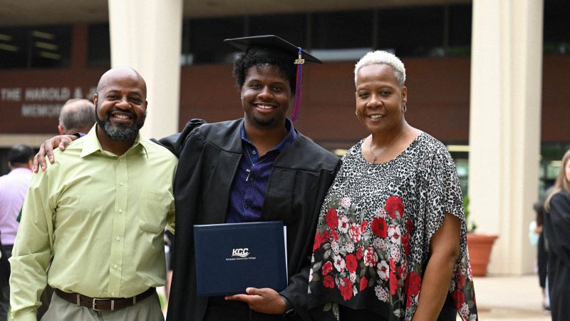 A student in Cap and gown with his parents from left to right in the front lawn of KCC.