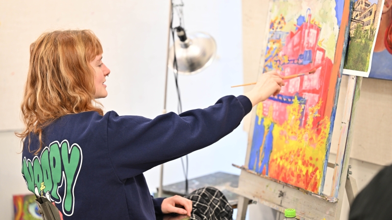 Student painting a bright, colorful architectural canvas on an easel in an art studio.